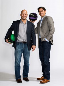 Anytime Fitness co-founders Dave Mortensen and Chuck Runyon standing and smiling with medicine balls wearing business casual attire. View larger image.