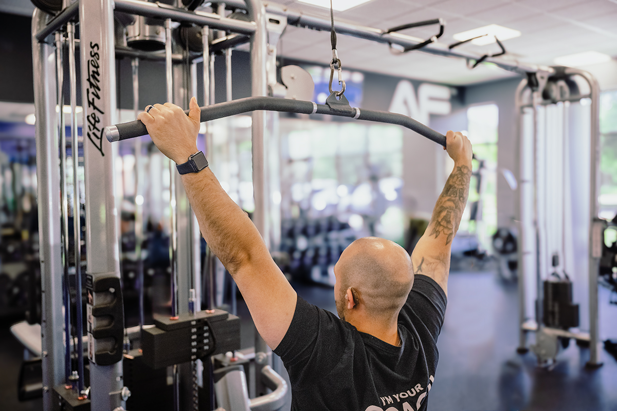 Close-up of Coach Mike demonstrating a wide grip on the lat pulldown bar in a gym.