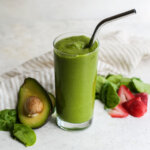 Glass of green smoothie sitting next to half an avocado and sliced strawberries.