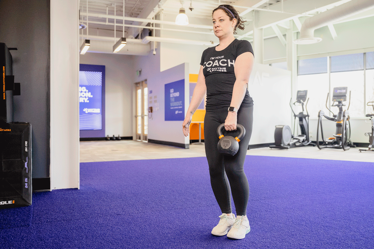 Coach Heather demonstrating a single-leg dumbbell Romanian deadlift in a gym setting.