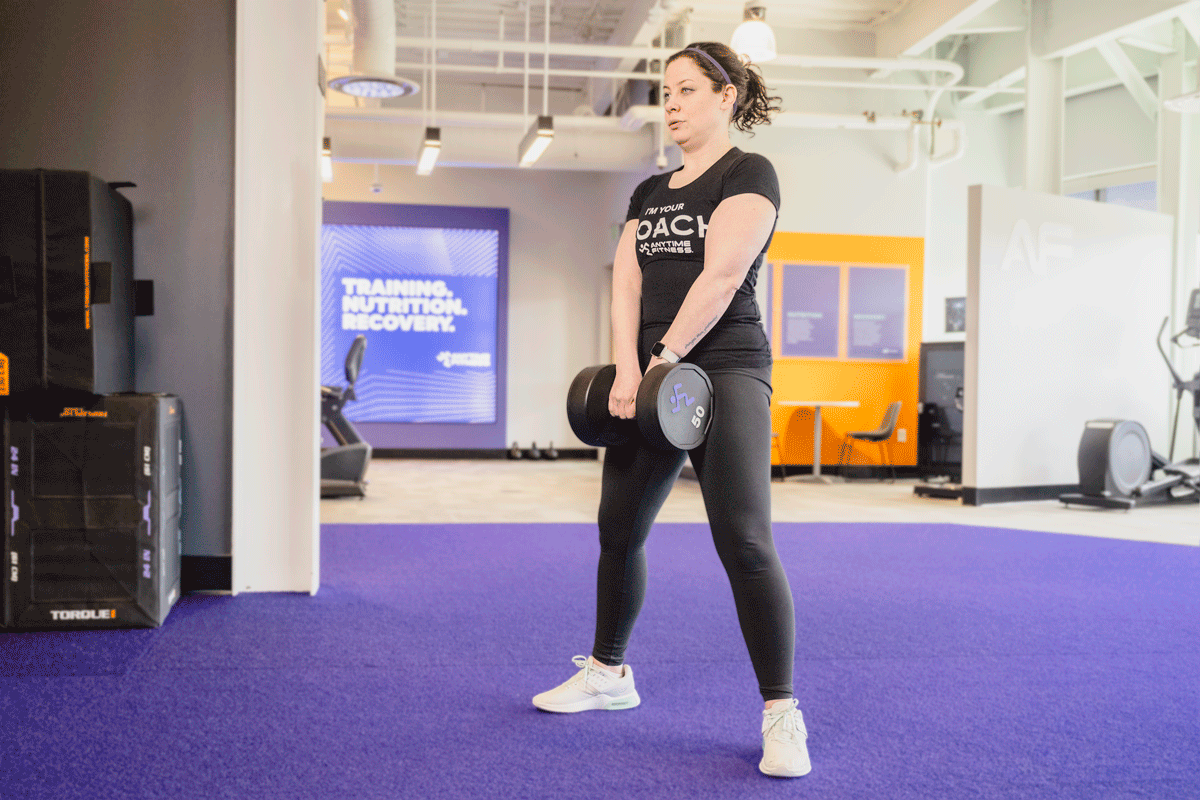 Coach Heather demonstrating a sumo dumbbell deadlift in a gym setting.