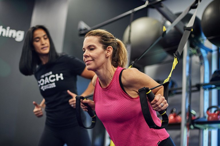 Anytime Fitness Coach training woman on TRX straps in a gym setting.