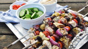Chicken and vegetable kabobs on a tray with ramekins of cucumbers, tomatoes, and hummus.