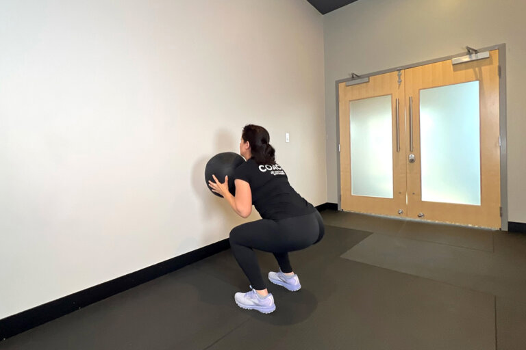 Coach squats in front of wall, holding medicine ball to chest.