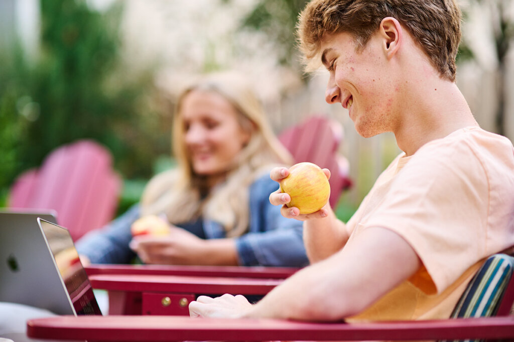 Man snacking on an apple while working on a laptop and sitting in a lawn chair in an outdoor setting.