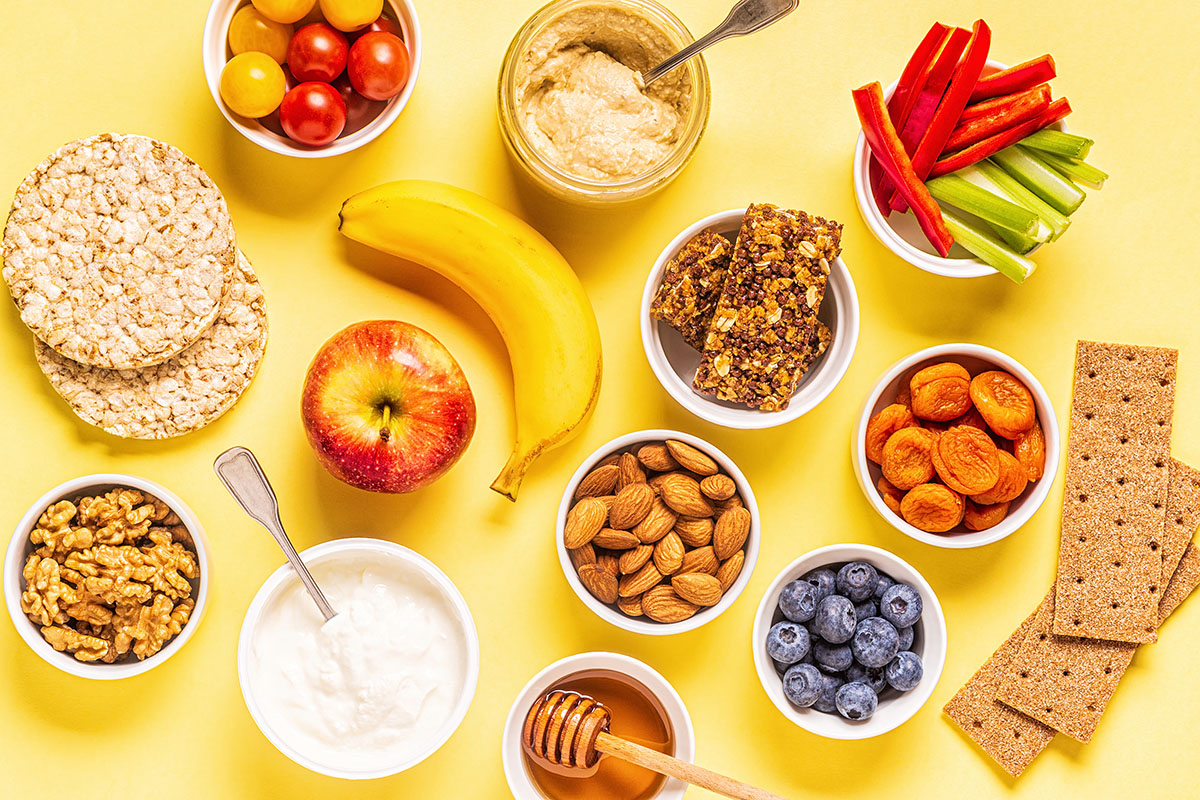 A variety of healthy grab-and-go snacks on a yellow background, including an apple, banana, blueberries, almonds, dried apricots, peppers, honey, rice cakes, and nuts.