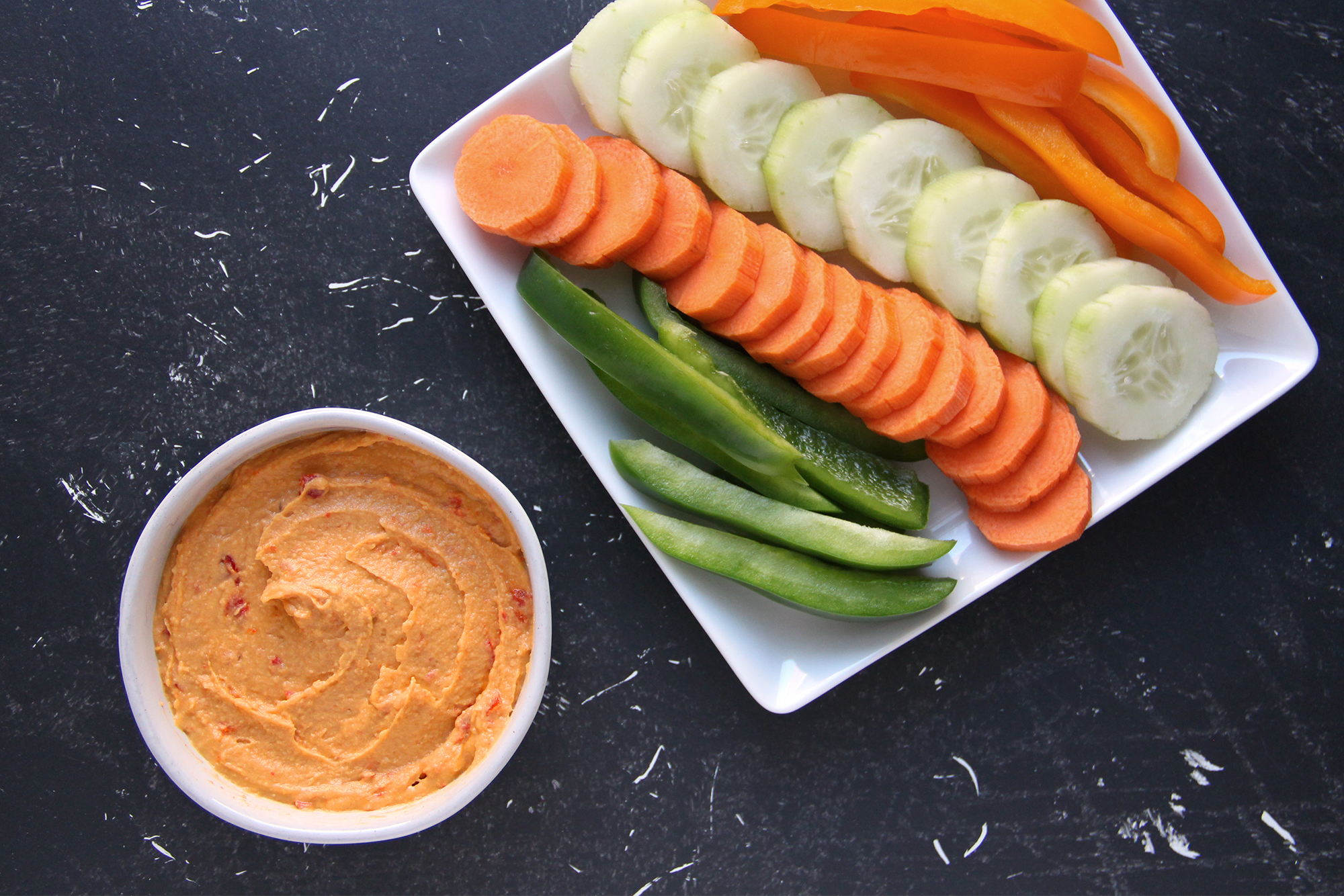 Plate of raw cut vegetables like carrots, cucumber, and bell pepper, next to a bowl of hummus.