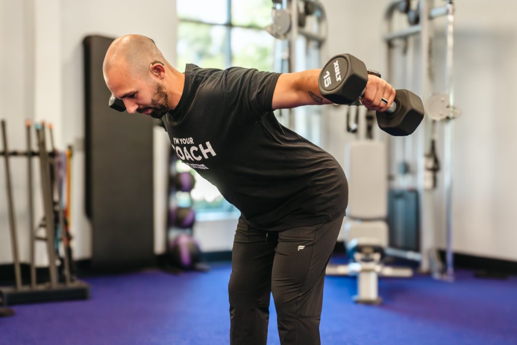 Anytime Fitness coach performing bent over lateral raise exercise with dumbbells, demonstrating proper form for shoulder and upper back muscle engagement.