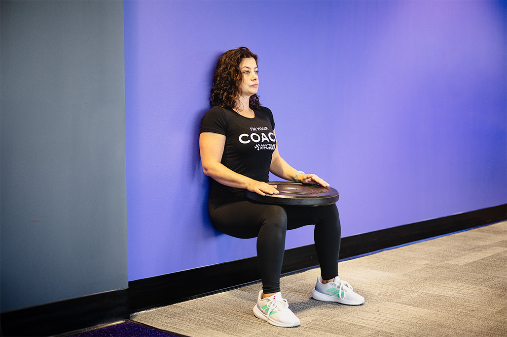 Woman performing a plate-resisted wall sit to increase lower body strength and endurance, targeting the quadriceps, glutes, and core muscles during this strength training exercise.