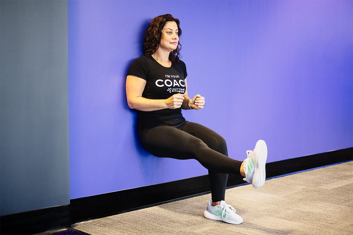 Female coach performing a challenging single-leg wall sit, demonstrating an advanced variation of the wall sit exercise for greater lower body strength and balance.