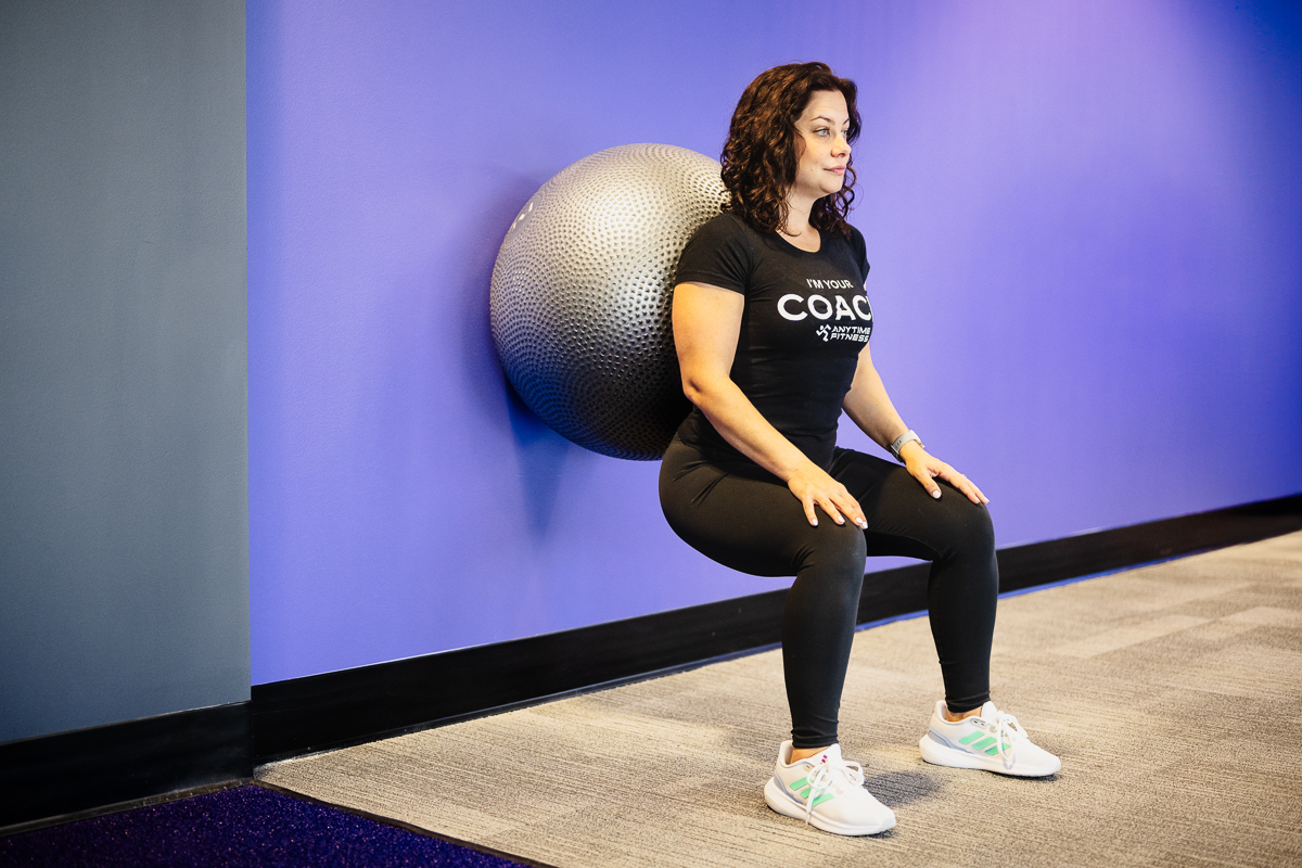 Animated GIF of a woman performing a stability ball wall squat, focusing on building lower body strength and improving balance with proper form in a gym setting.