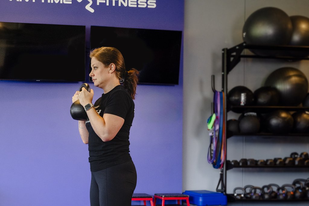 A close-up image of a woman performing a goblet squat while holding a kettlebell at chest level. The gym setting includes a purple wall with the "Anytime Fitness" logo and various fitness equipment in the background.