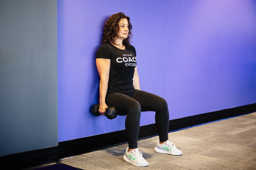 Woman performing a wall sit with dumbbells in a gym setting, emphasizing lower-body strength and endurance.