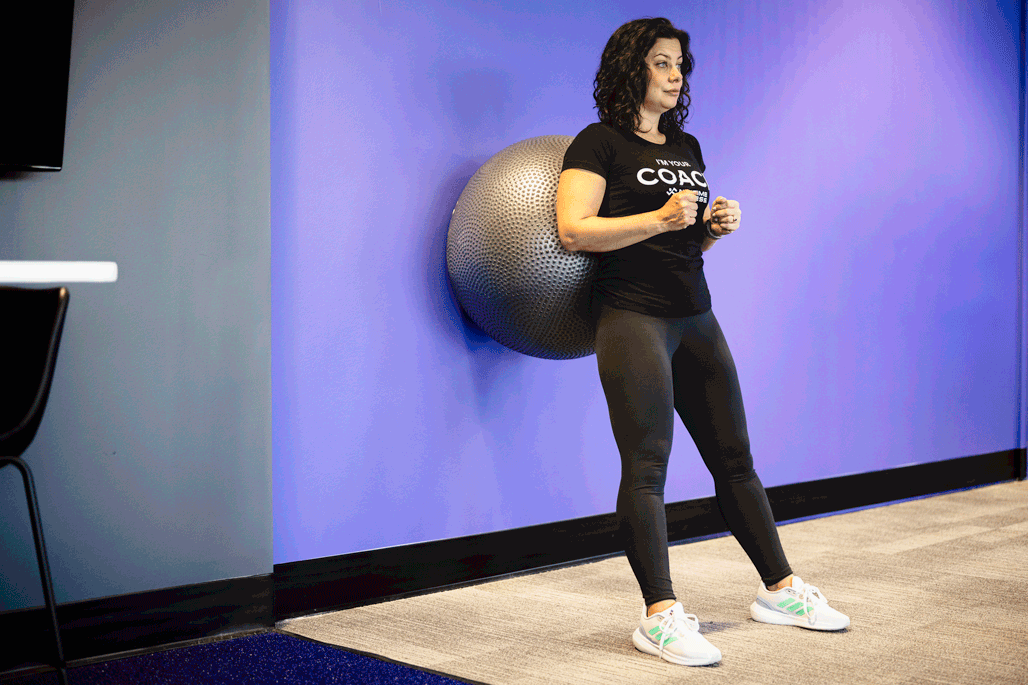 Woman performing a wall squat with a stability ball for added support. This exercise targets lower body muscles like the quads and glutes, while improving balance and posture in a gym environment.