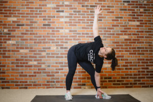 Anytime Fitness Coach performing a windmill exercise on a fitness mat in a home gym.