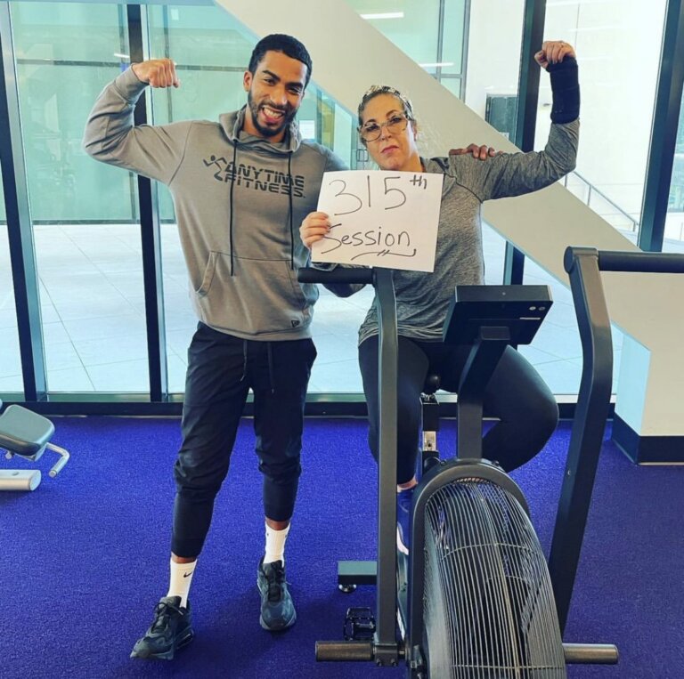 Coach and his client in a gym, both flexing and smiling, the client is holding a sign that says “315th session.”
