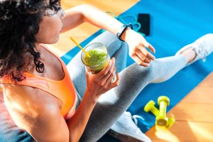 Woman in workout clothes drinking a green smoothie while checking her fitness tracker after a workout on a blue exercise mat with dumbbells and a smartphone nearby.