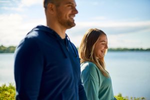 A smiling couple enjoying a walk near a lake on a sunny day, highlighting the health benefits of walking outdoors for mental well-being.