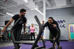 An Anytime Fitness Coach works with a woman pushing a sled in a gym.