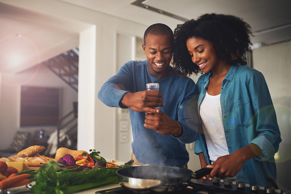 A couple is happily cooking together. The man is wearing a blue sweater and pouring seasoning into a pan, and the woman next to him is wearing a blue shirt while holding the pan on the stove.