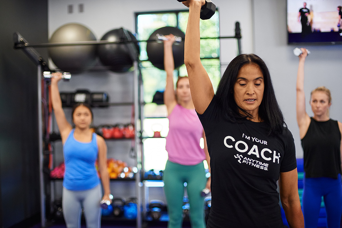 An Anytime Fitness Coach leads a group strength training session, working the upper arms with dumbbells.