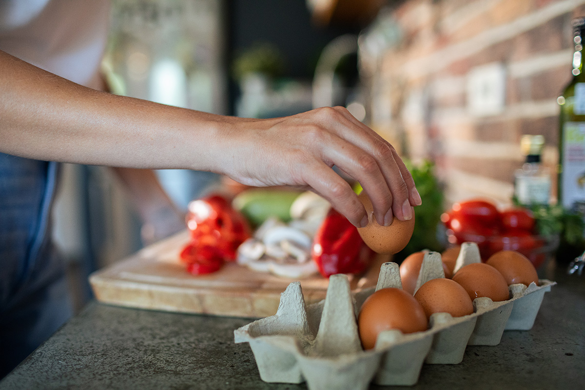 Someone is holding an egg in their hand over a carton of eggs next to a cutting board with fresh vegetables on it.