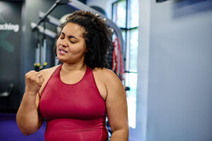 A woman in a red tank top confidently flexing her arm at the gym, symbolizing strength and personal fitness progress.