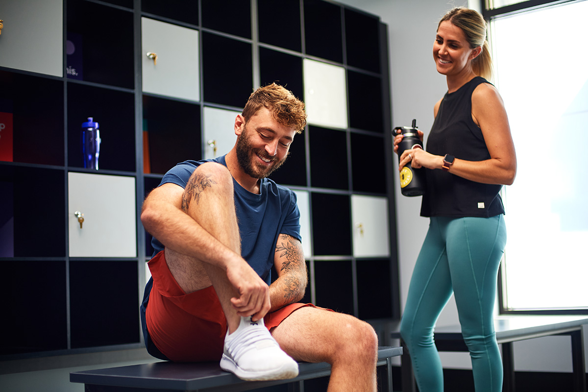 A man sitting on a bench tying his shoes while smiling and chatting with a woman holding a water bottle in a gym locker room.