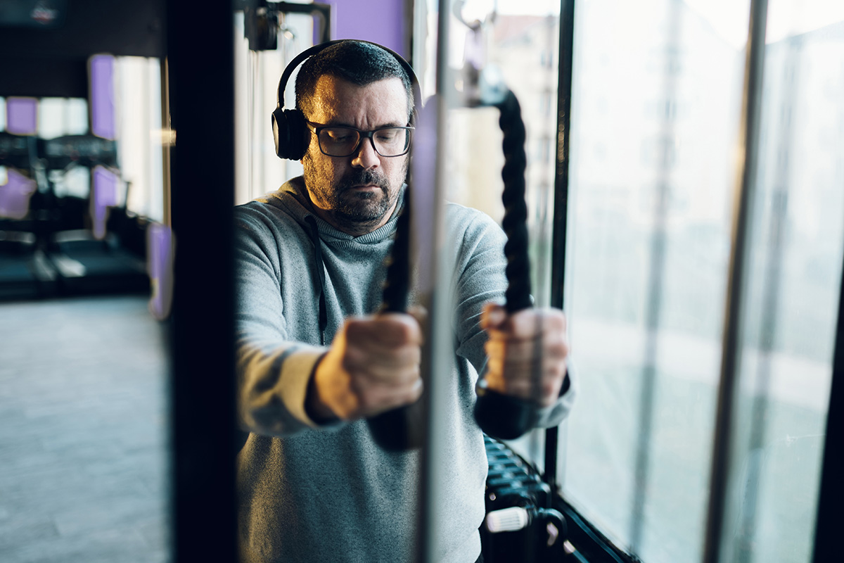 A man wearing headphones and a hoodie, performing a cable machine exercise at the gym with focus and determination.