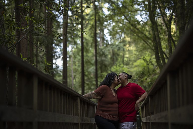 Candie and her wife, Lorena, smiling and embracing while standing on a bridge in a scenic forest.