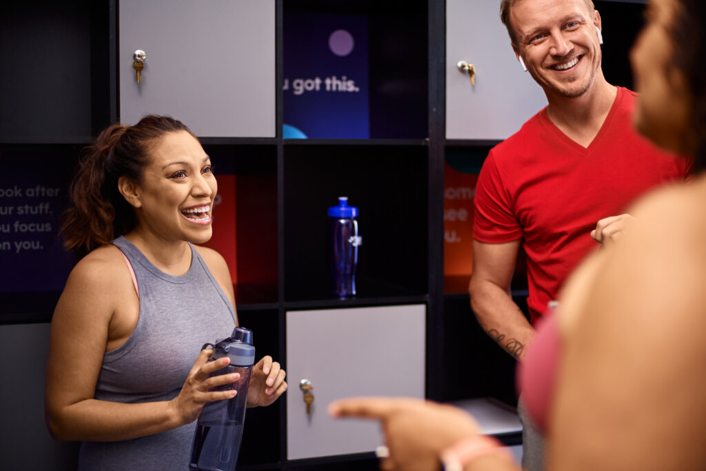 Woman smiling in the gym lobby near lockers, with her friends nearby smiling and conversing.