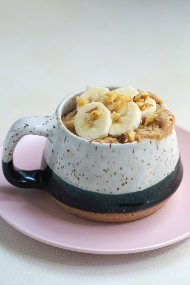 Side view of a banana bread in a mug with sliced bananas and walnut topping on a pink plate.