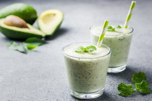 Close-up image of two green smoothies with green straws, made with avocado, mint, spinach, banana, and chia seeds.