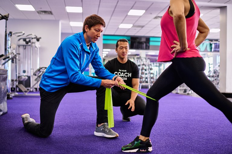 A coach and a physical therapist help a woman stretch her legs in a gym using a resistance band.