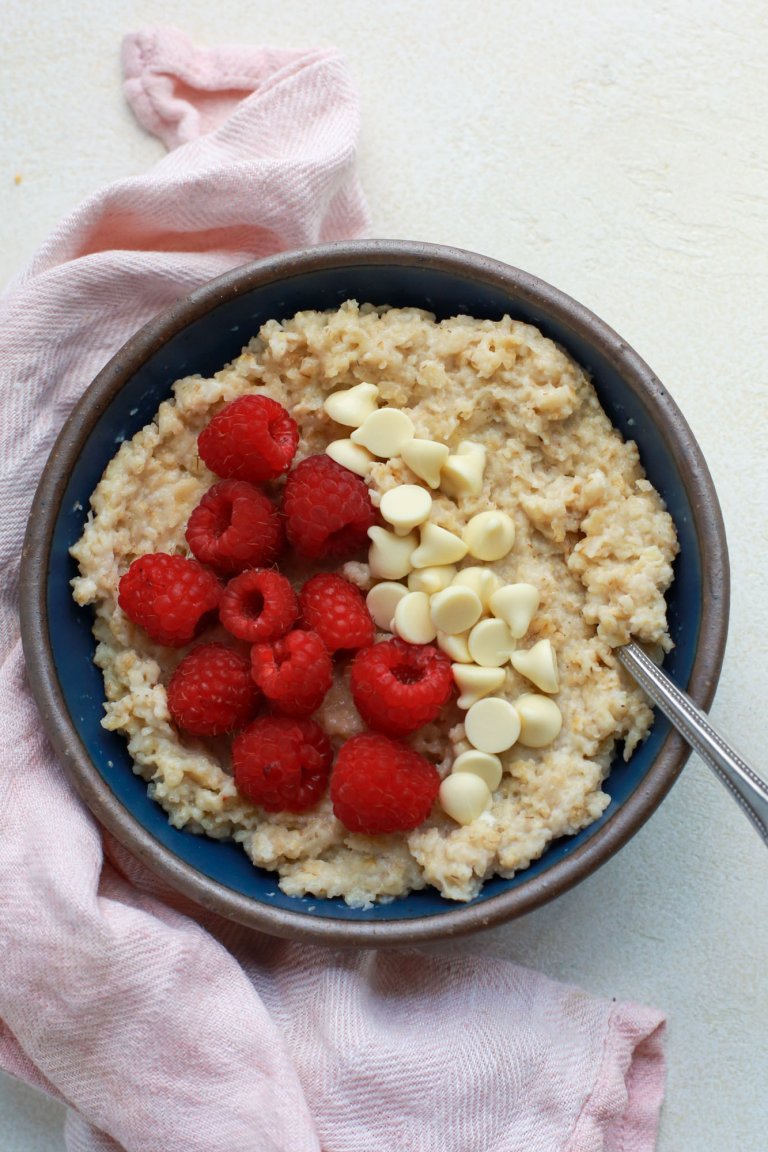 Bowl of oats prepared with raspberries and white chocolate chips as toppings. shown ready-to-eat with a spoon.