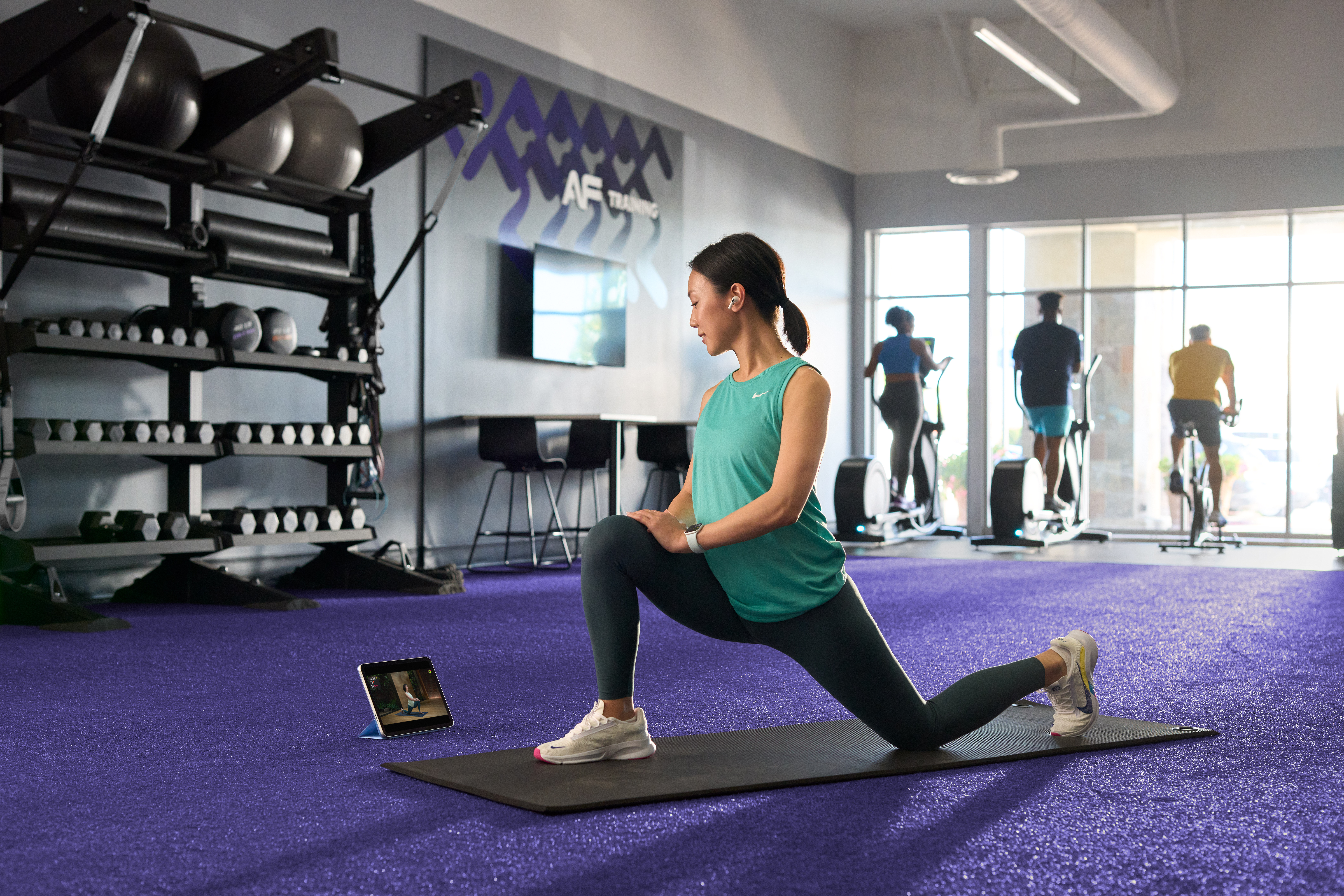 A woman in a blue tank top does an Apple Fitness+ yoga session on a mat in an Anytime Fitness gym.