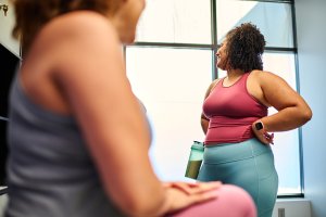 Two women in athletic wear chatting in a gym, one holding a water bottle, emphasizing fitness and hydration.