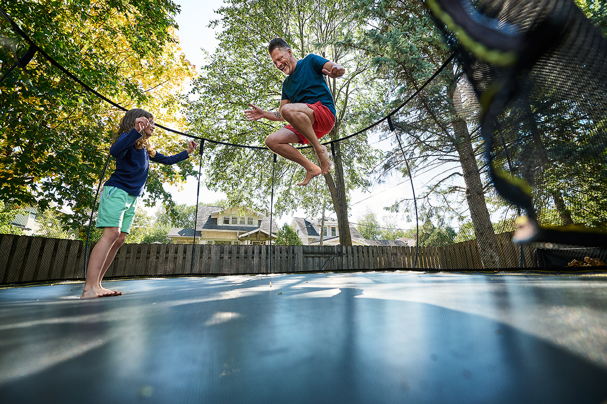 Father and child jumping on a trampoline in a backyard, enjoying outdoor playtime under the trees