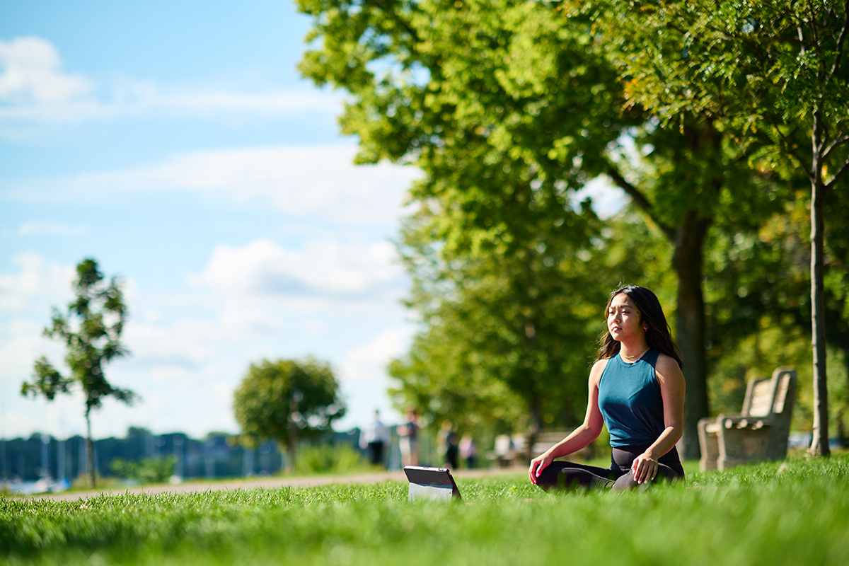 Woman meditating in a park on a sunny day, surrounded by greenery and blue skies.