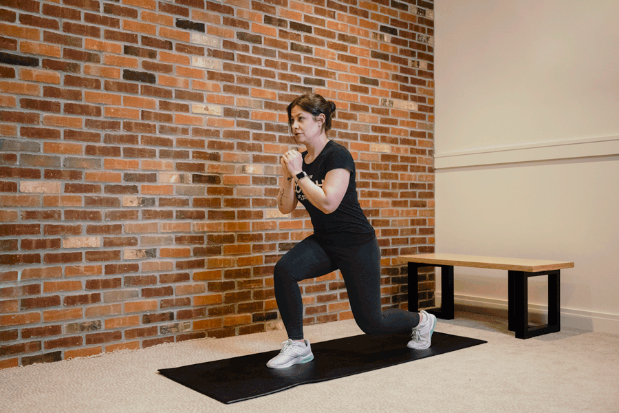Coach Heather demonstrating an alternating jump lunge on an exercise mat with a bench and a brick wall behind her.