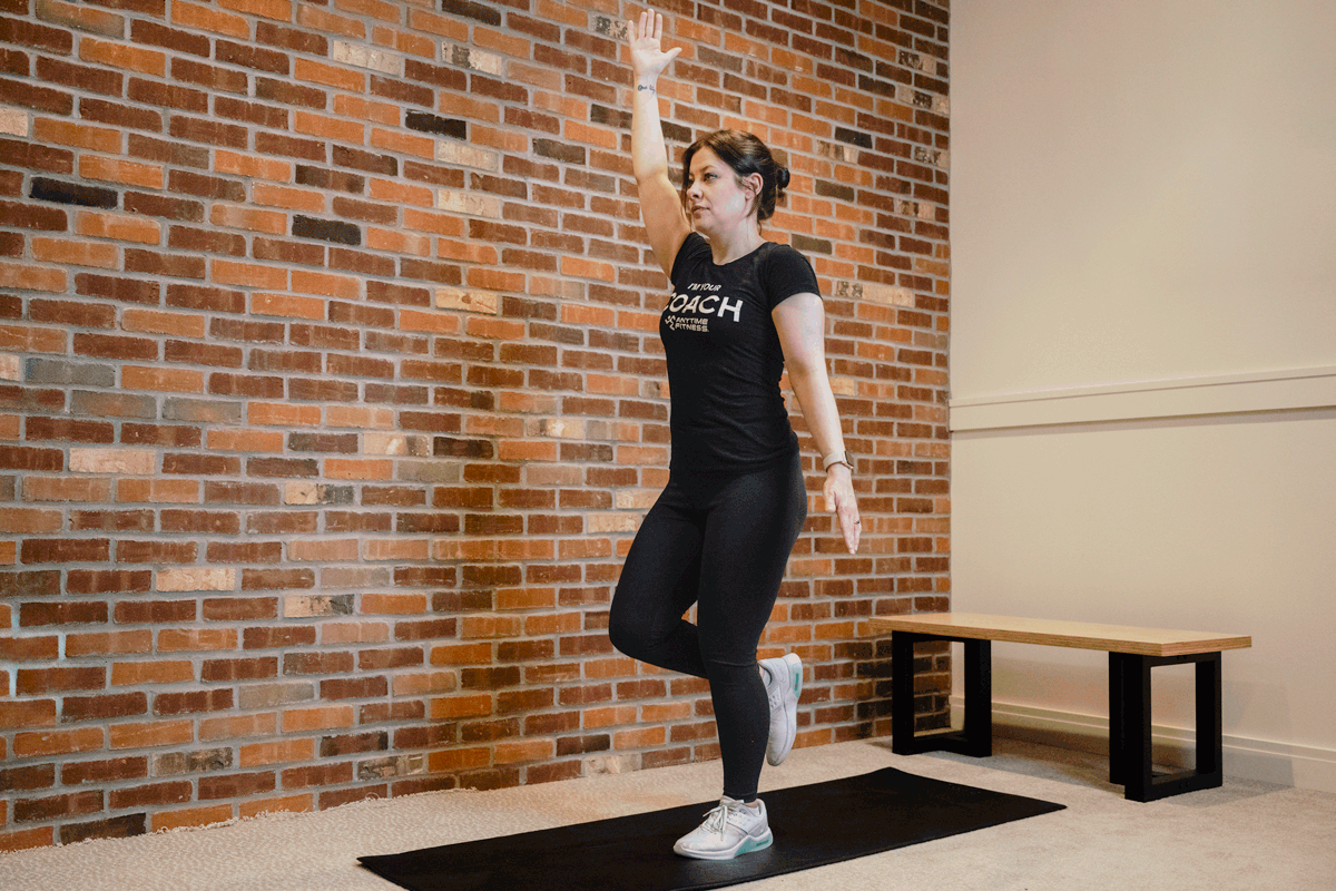 Coach Heather demonstrating a single-leg squat reach on an exercise mat with a bench and a brick wall behind her.