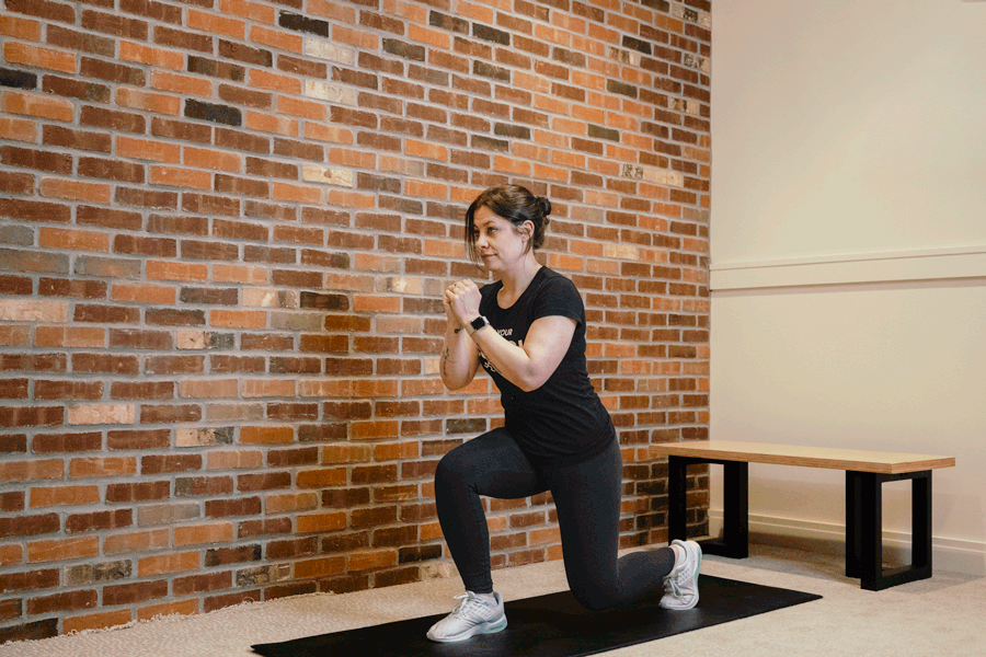 Coach Heather demonstrating split jumps on an exercise mat with a bench and a brick wall behind her.
