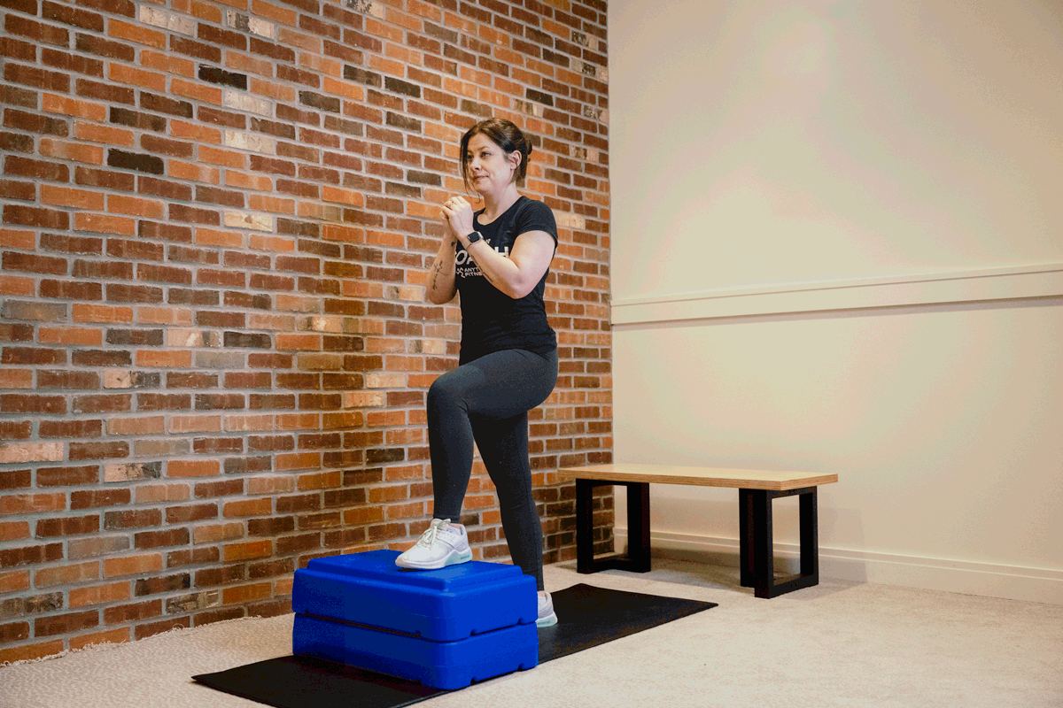 Coach Heather demonstrating step-ups on an exercise mat with a bench and a brick wall behind her.