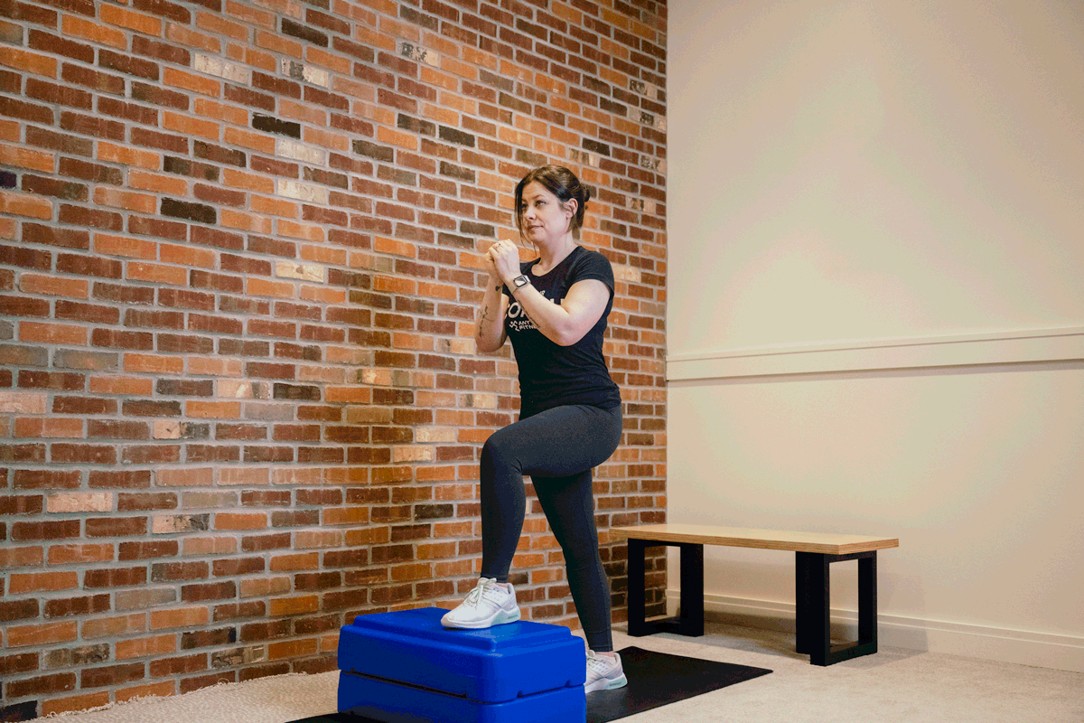 Coach Heather demonstrating step-ups to knee drive on an exercise mat with a bench and a brick wall behind her.