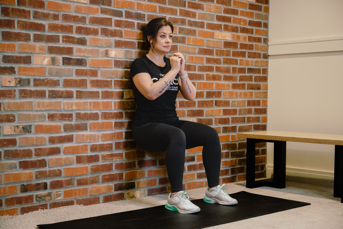 Coach Heather demonstrating a wall sit with a bench and a brick wall behind her.