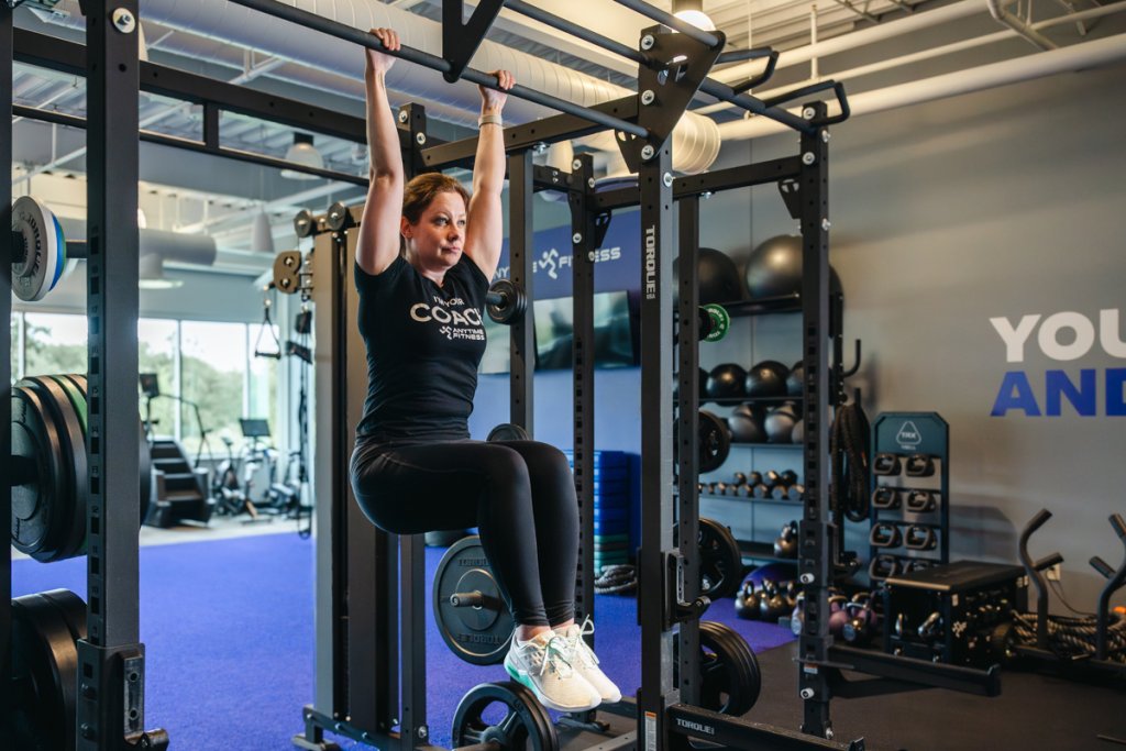 Personal trainer executing a hanging knee raise exercise, engaging core and lower abs. The image shows the trainer in mid-movement with knees raised.