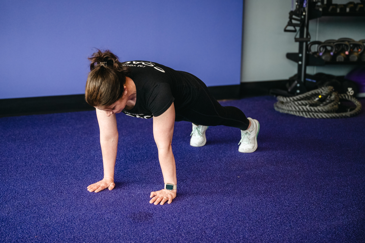 Anytime Fitness coach in a high plank position on a blue gym floor, showing a core strengthening exercise as part of a treadmill warmup routine.