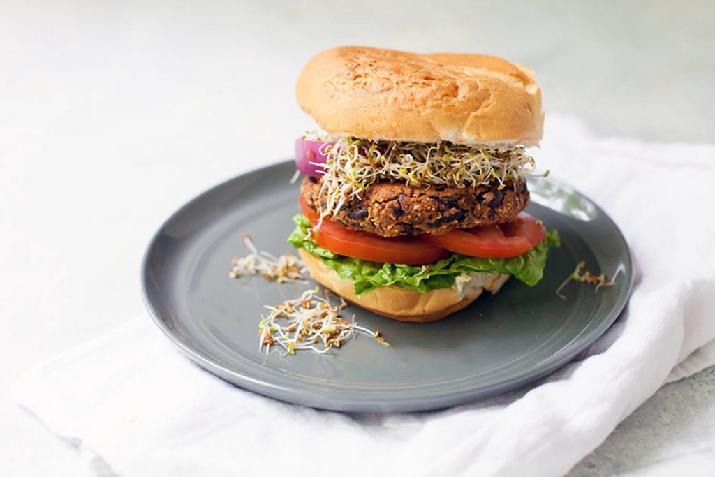 A hearty black bean burger topped with fresh lettuce, tomato slices, red onion, and alfalfa sprouts, served on a toasted bun. The burger is presented on a gray plate with a white napkin.