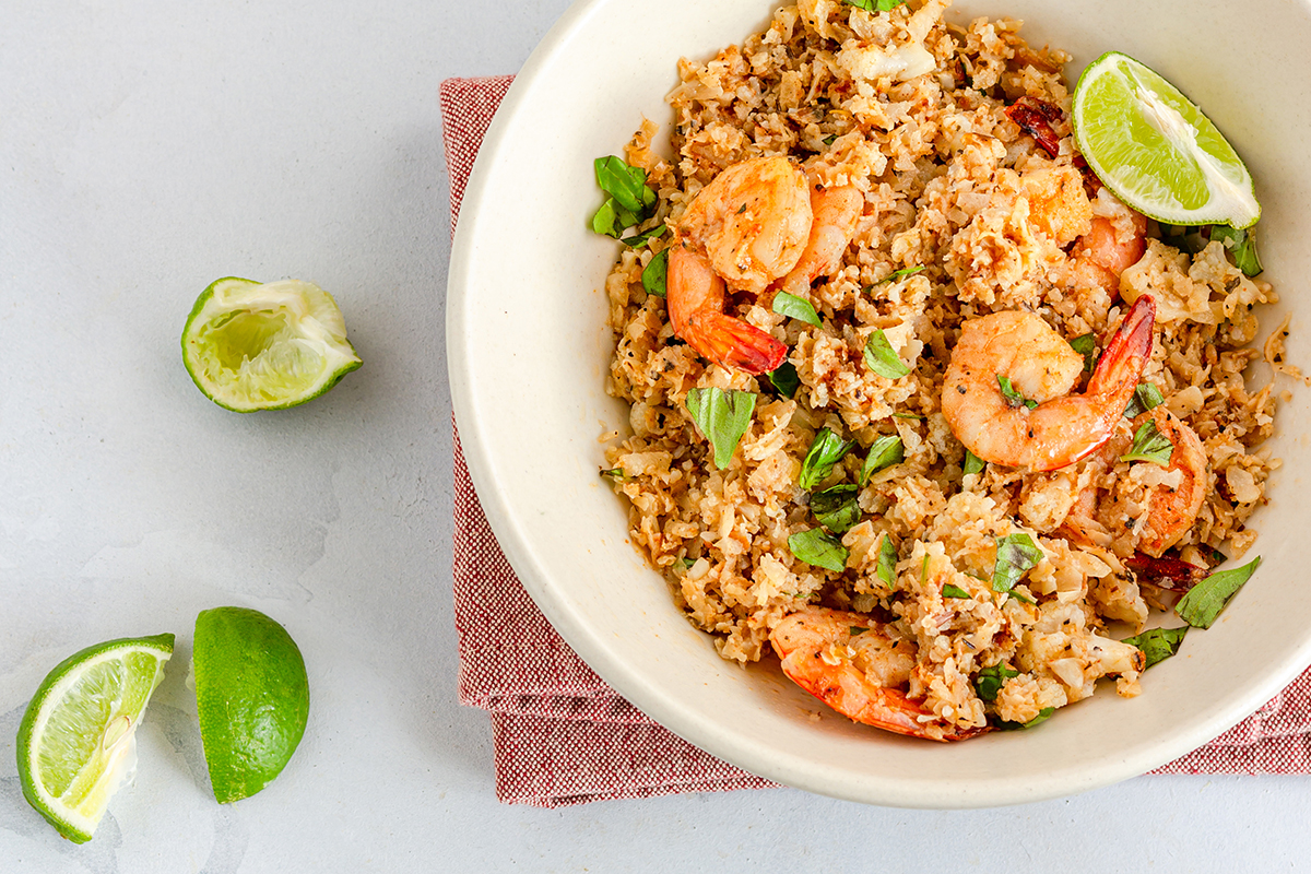 A bowl of shrimp cauliflower fried rice garnished with fresh basil and lime wedges, accompanied by a pair of red chopsticks. The dish is served on a light background with scattered cauliflower florets and basil leaves.