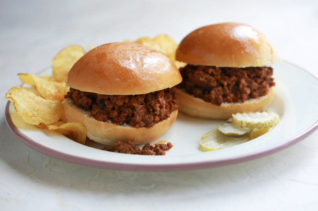 Two classic sloppy joes with rich, savory meat filling on soft buns, served with crispy potato chips and sliced pickles on a white plate.